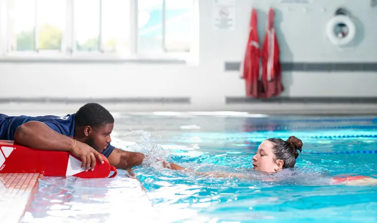 Lifeguard lies down on the edge of an indoor pool and reaches out to assist a swimmer